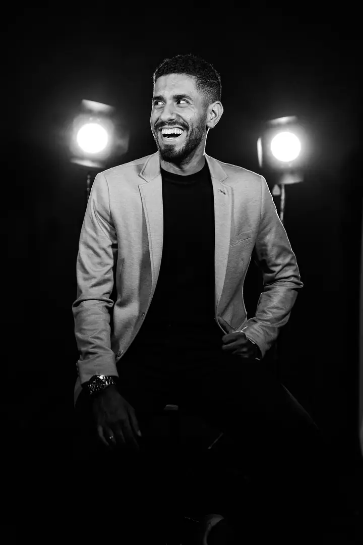 Natanael Olan sorrindo, sentado entre dois refletores de estúdio